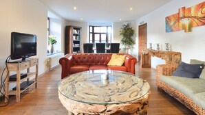 Ein Wohnzimmer mit einer Ledersofa, einem Glastisch, einem Fernseher auf einem Holzboden, einem Bücherregal und einem Essbereich im Hintergrund.