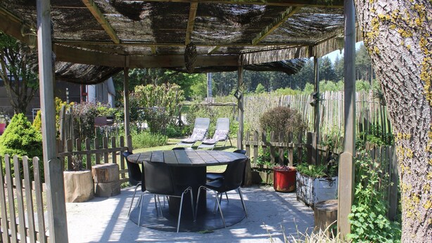 Eine überdachte Terrasse im Freien mit einem runden Tisch und Stühlen, umgeben von einem Holzzaun und Pflanzkübeln.