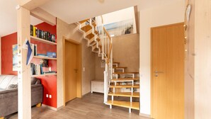 A modern interior with a wooden staircase, a red wall with bookshelves, a grey sofa, and a wooden door. - Modernes Ferienhaus mit Sauna in Wiek am Meer (Wiek)