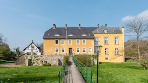 A yellow building with a traditional design, a stone wall, and a green pathway leading to the entrance. - Spacious Apartment in Leopoldshöhe near Teutoburg Forest (Leopoldshoehe)