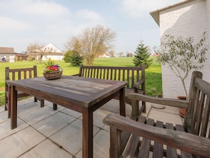Une terrasse extérieure avec une table et des chaises en bois, un panier en osier rempli de fleurs et une vue sur un champ verdoyant et des maisons au loin.