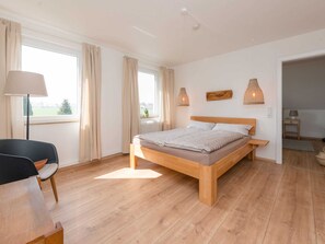 Une chambre à coucher avec un lit en bois, un bureau avec une chaise et une vue sur l’extérieur grâce à de grandes fenêtres.