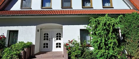 A two-story building with a red-tiled roof, white exterior walls, and a central entrance with a door and steps.