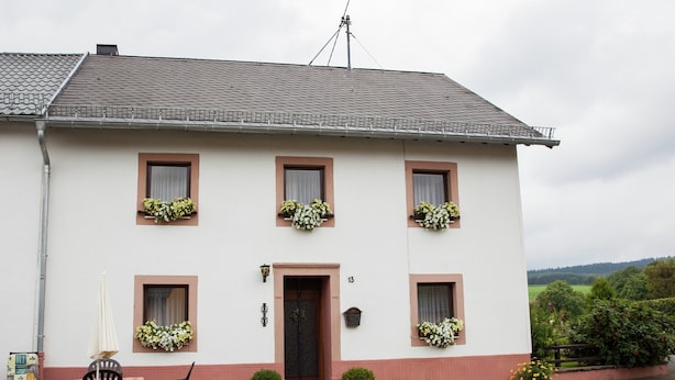 Un bâtiment blanc avec une base rouge, doté de six fenêtres ornées de jardinières, d’une porte et d’une cour dallée avec des chaises et des tables.