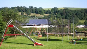 Un terrain de jeux avec des balançoires et un toboggan, situé dans un espace verdoyant, avec une maison et des arbres en arrière-plan.