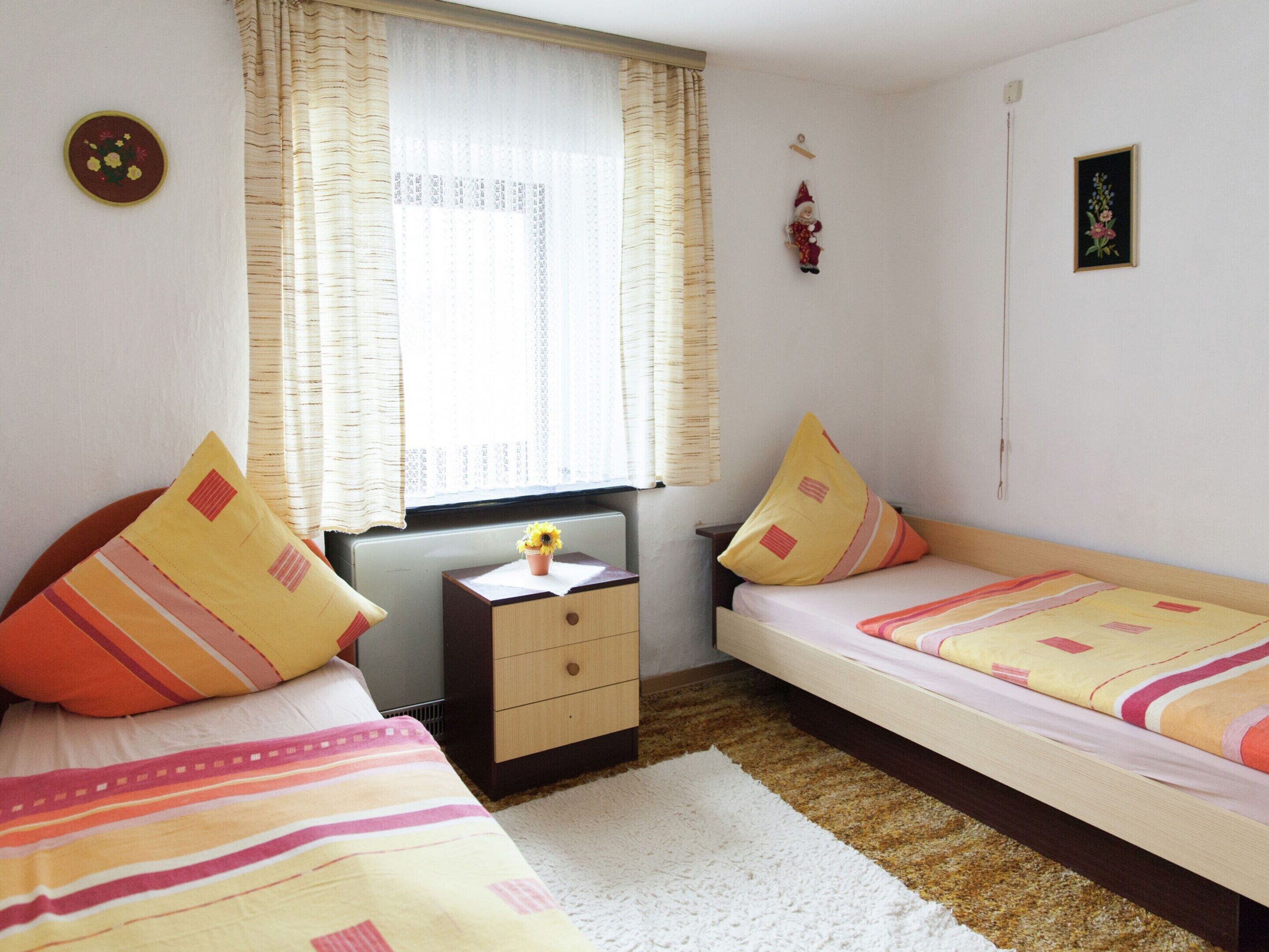 A small, well-lit bedroom with two single beds, a nightstand, a radiator, and a window with curtains.