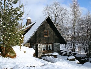 A rustic cabin with a stone foundation, wooden walls, and a steeply pitched roof, surrounded by snow-covered trees and a wooden fence.