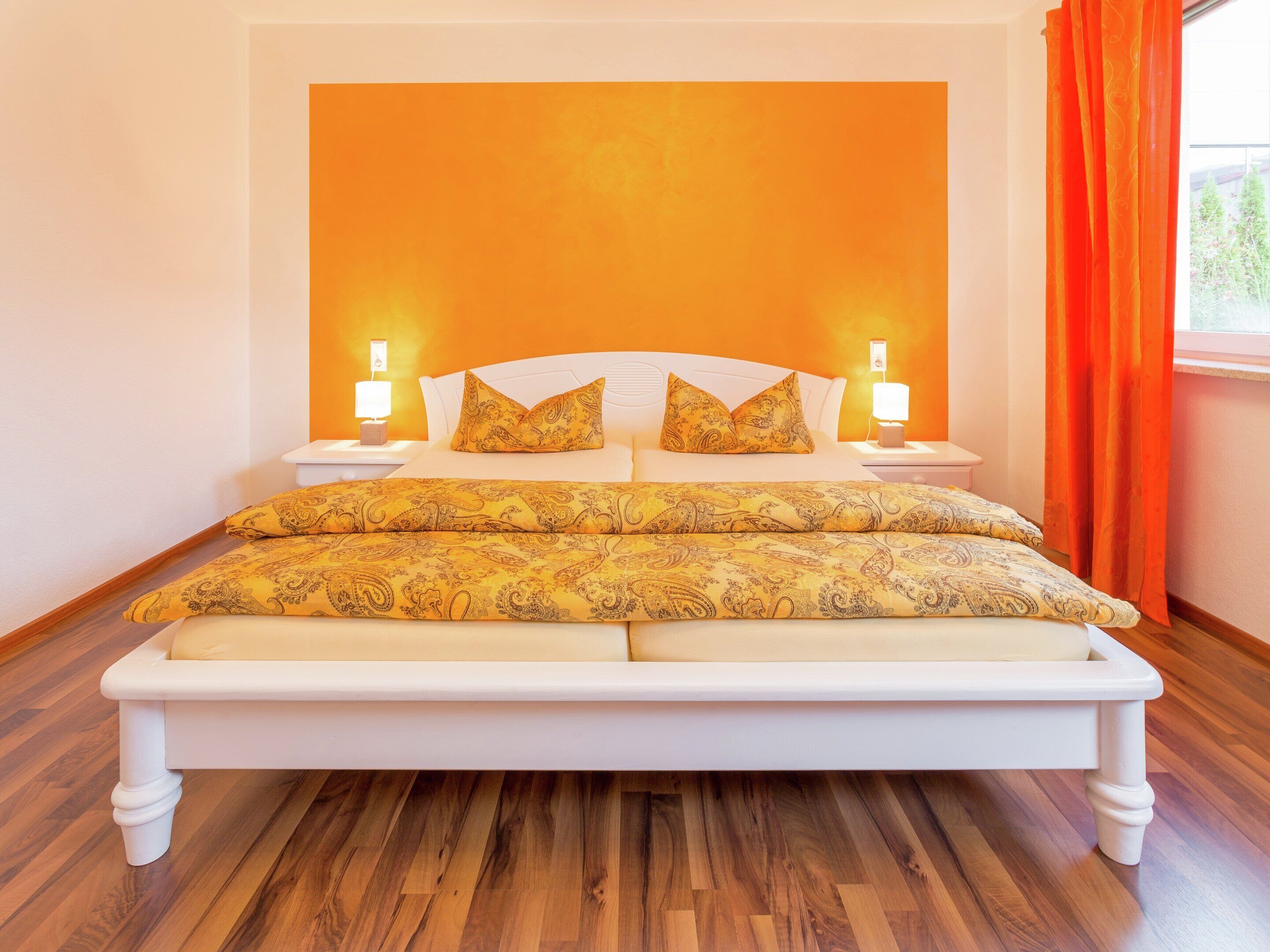 Quarto com cama grande, duas luminárias de cabeceira e janela com cortinas laranja.