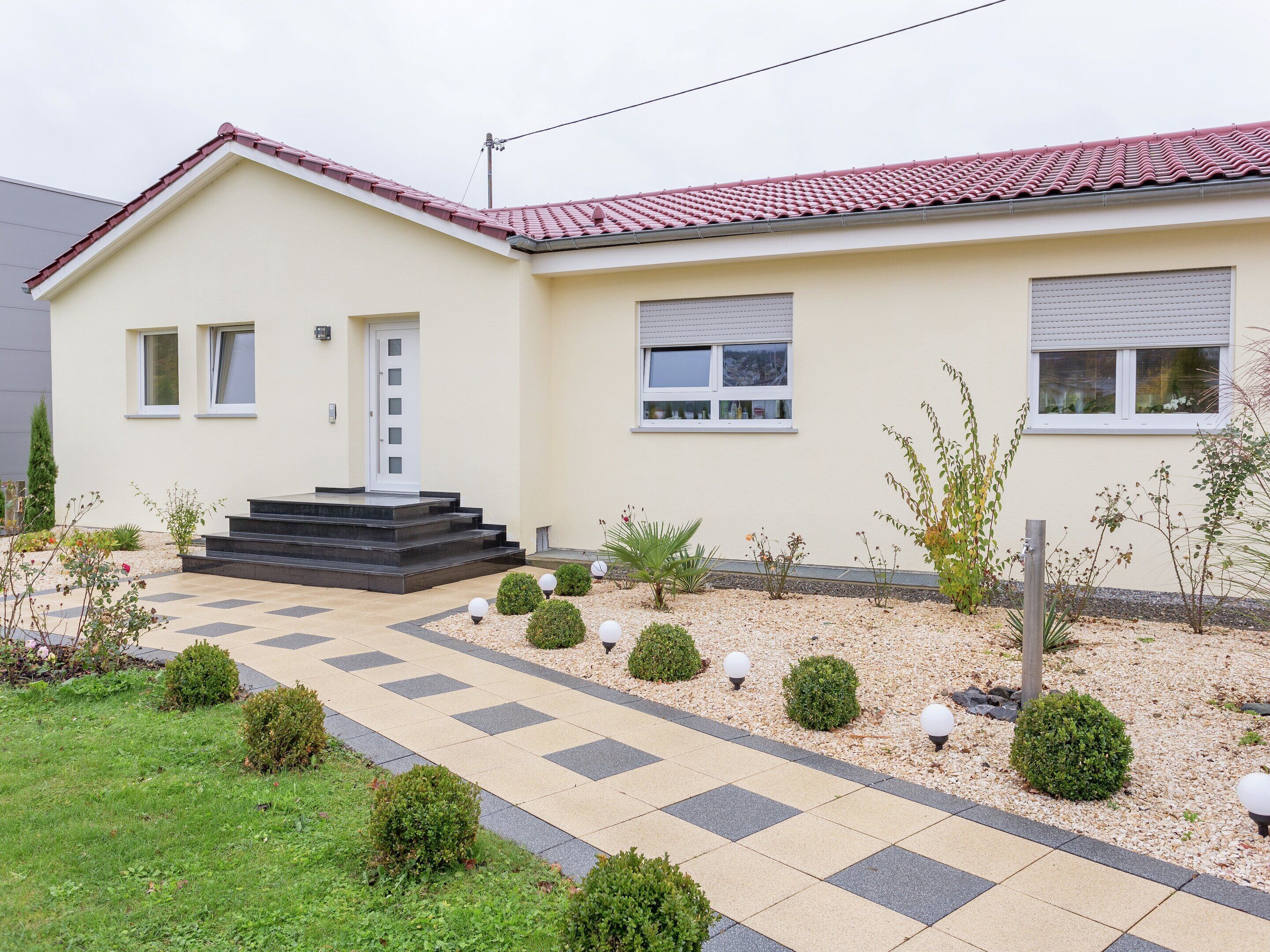 A modern two-story house with a red-tiled roof, a front entrance with steps, and a garden with small bushes and decorative lights.