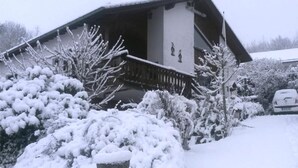 A residential building with a sloped roof, a wooden deck, and snow-covered bushes and trees.