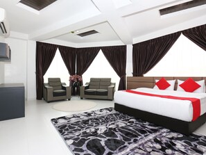 Quarto de hotel moderno com uma cama grande, duas poltronas, uma mesa pequena com um vaso de flores e vista da cidade através de amplas janelas.