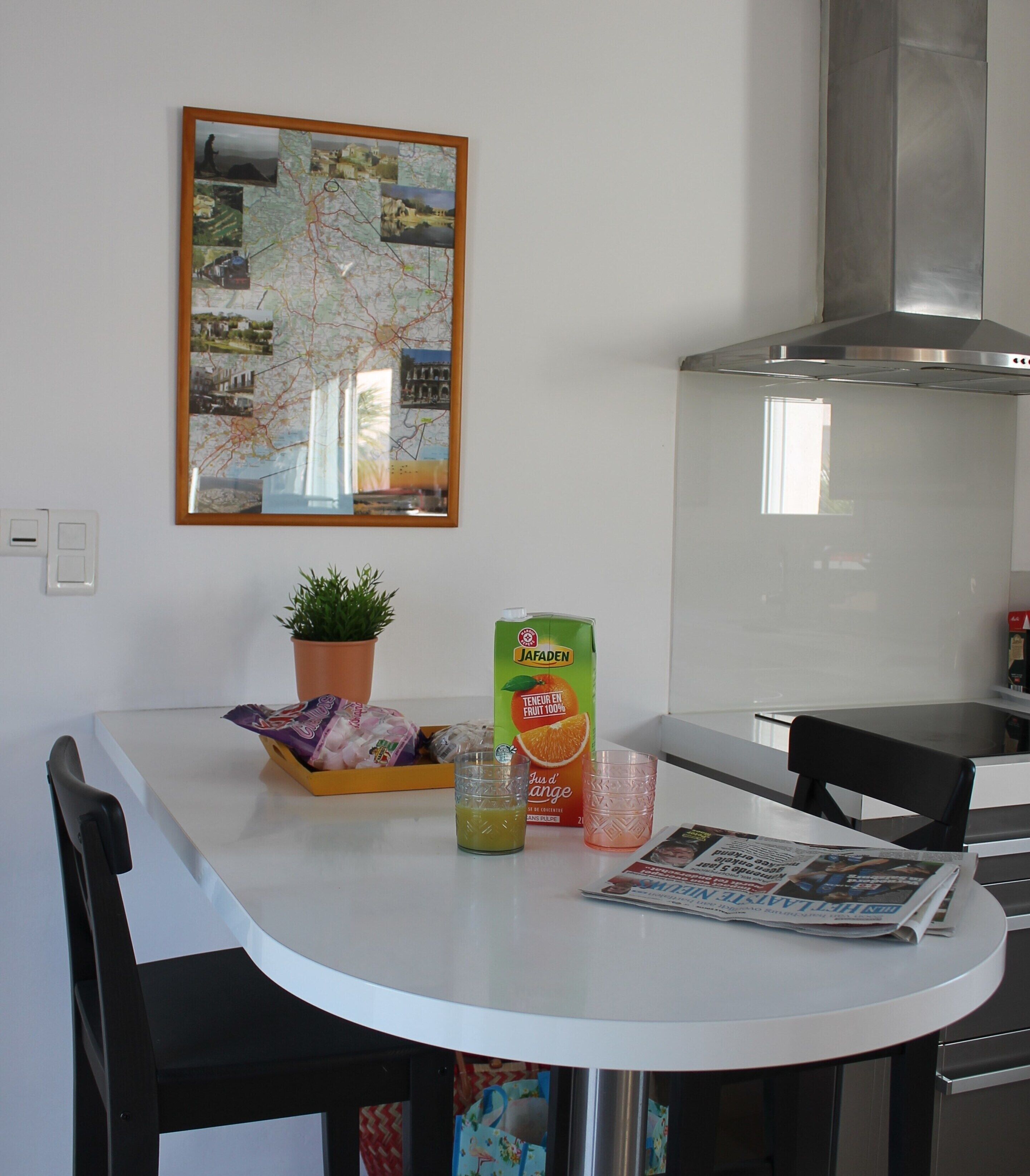 A kitchen with a white table, a potted plant, a carton of juice, and a newspaper.
