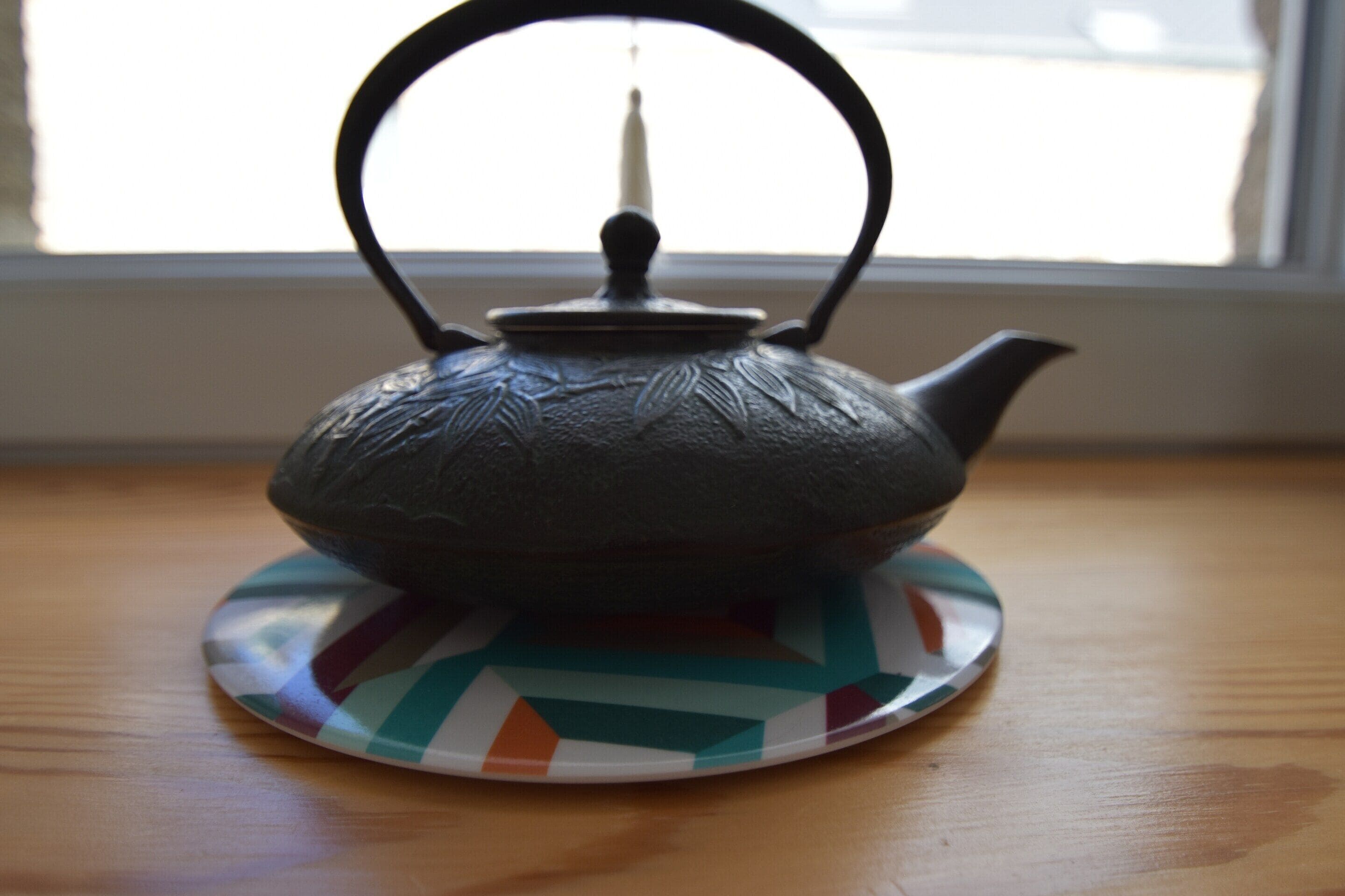 A teapot on a patterned coaster on a wooden table near a window.
