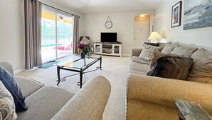 Un salon avec un canapé, une table basse et un téléviseur. On y trouve également un ventilateur de plafond, une horloge au mur et un tableau.
