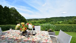 Un tavolo all'aperto con tovaglia floreale, una bottiglia di vino e bicchieri, con vista panoramica su colline verdi e cielo sereno.