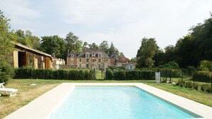 Una piscina rectangular con cubierta de madera, rodeada por un césped cuidado y un edificio de ladrillo grande al fondo.