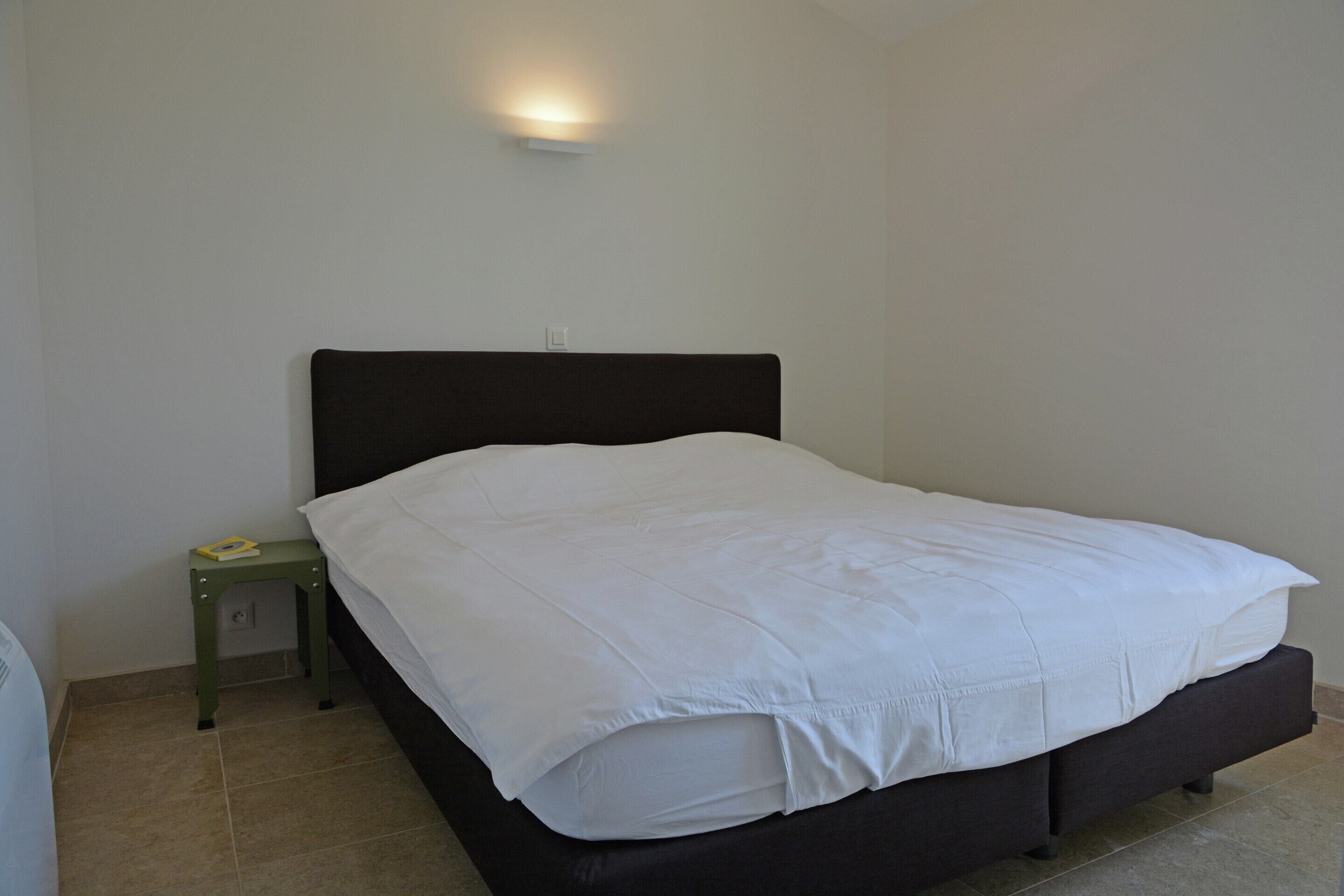 A bed with a dark headboard, a small bedside table with a book, and a wall-mounted light.