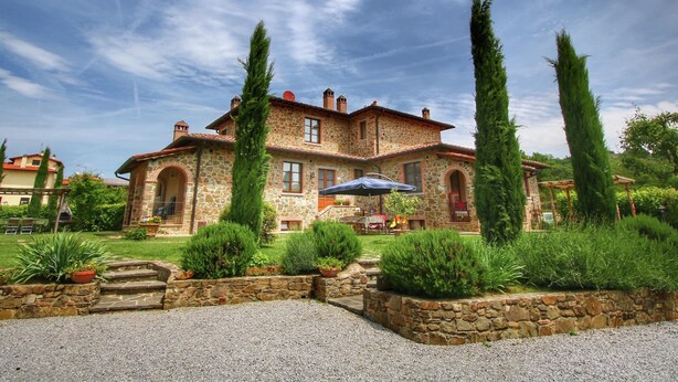 A stone house with a patio, surrounded by well-maintained gardens and tall trees. - Cottage in Tuscany With Pool & Olive Groves (Lucignano)