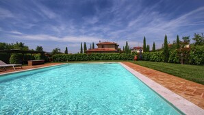 A rectangular swimming pool with clear blue water, surrounded by a brick patio. In the background, there is a building with a red-tiled roof and a garden with neatly trimmed hedges and tall trees. - Cottage in Tuscany With Pool & Olive Groves (Lucignano)