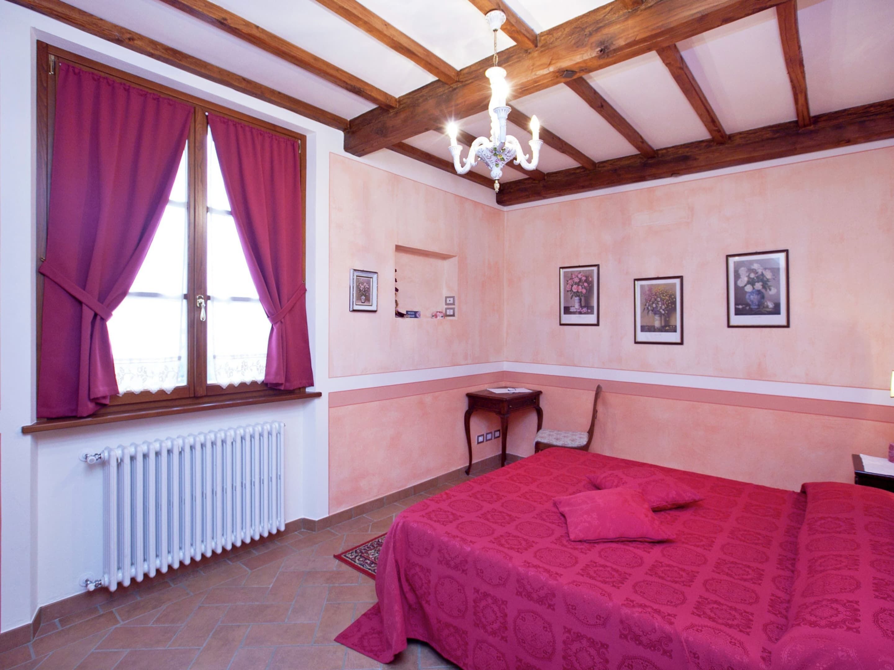 A bedroom with a bed, a window with curtains, a radiator, a wooden ceiling, and wall-mounted pictures.