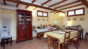 A dining area with a wooden table set for four, surrounded by chairs. A large wooden cabinet with glass doors is to the left. The room has exposed wooden beams on the ceiling and tiled flooring. - Cottage in Tuscany With Pool & Olive Groves (Lucignano)