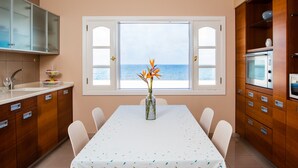 Eine Küche mit Esstisch, Stühlen, einer Blumenvase, Meerblick durch ein Fenster und Holzschränken.