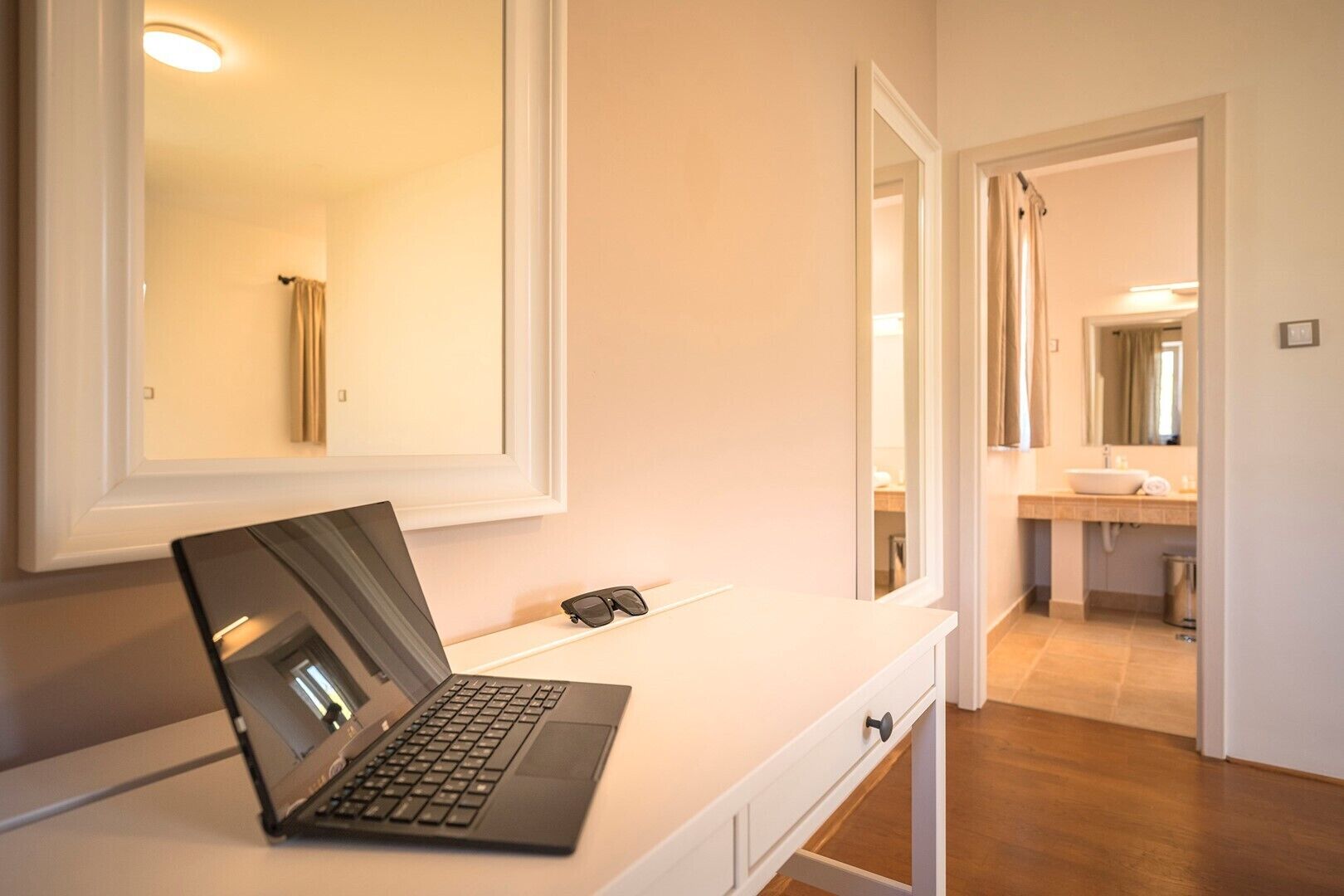 A modern bathroom with a white vanity, a laptop, and a pair of sunglasses on the counter.