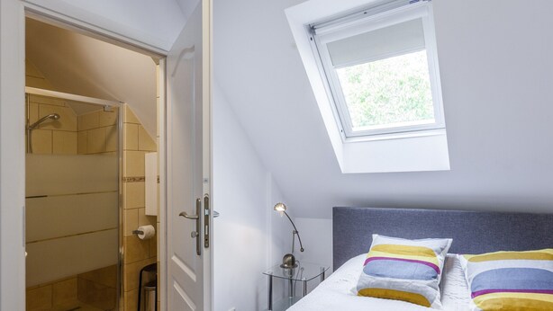 Un dormitorio con tragaluz, una cama con almohadas de colores, una mesita de noche con lámpara y una ducha a ras de suelo.