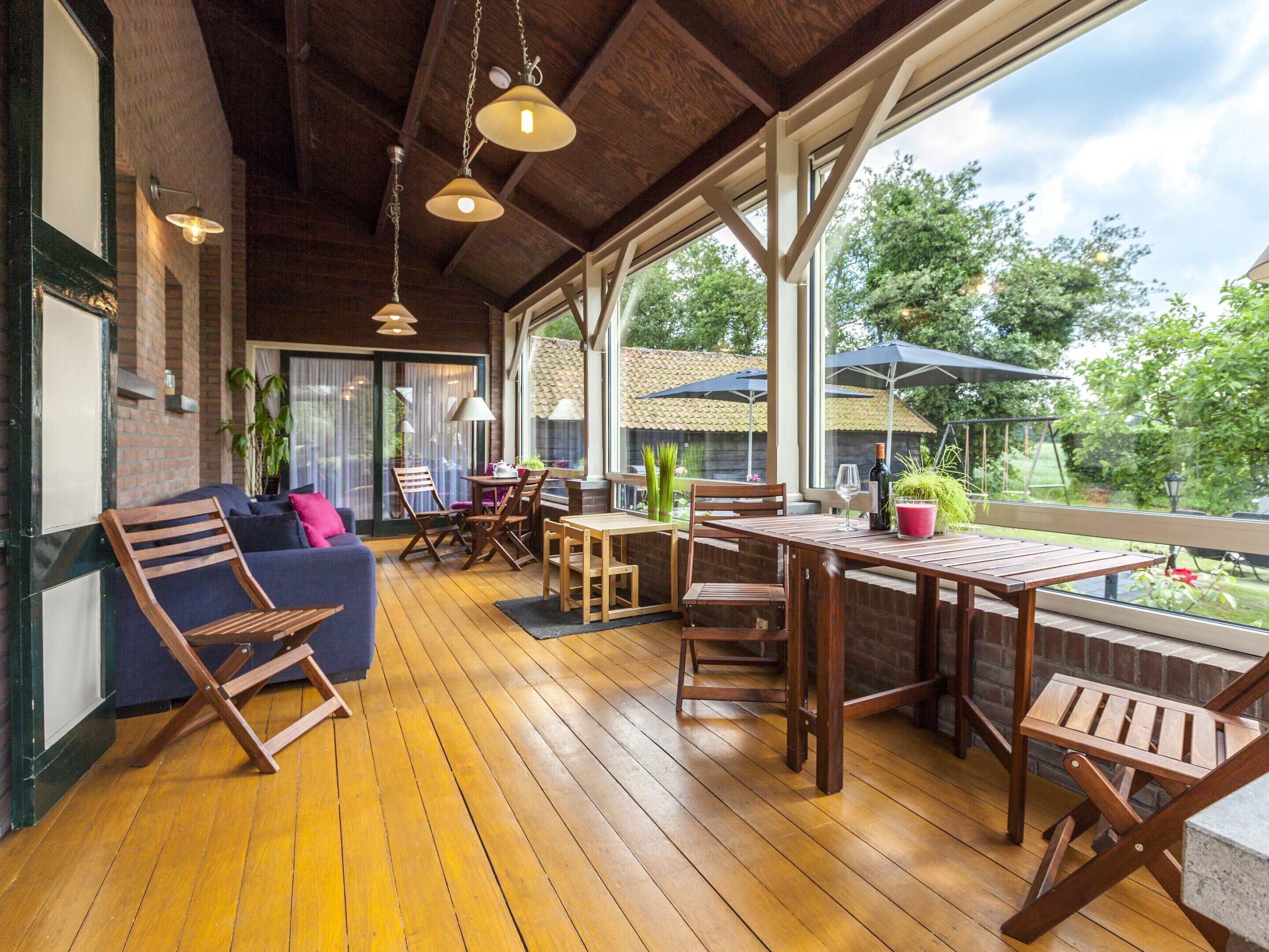A covered patio area with wooden flooring, a wooden table, and chairs. There are potted plants and a wine bottle on the table.