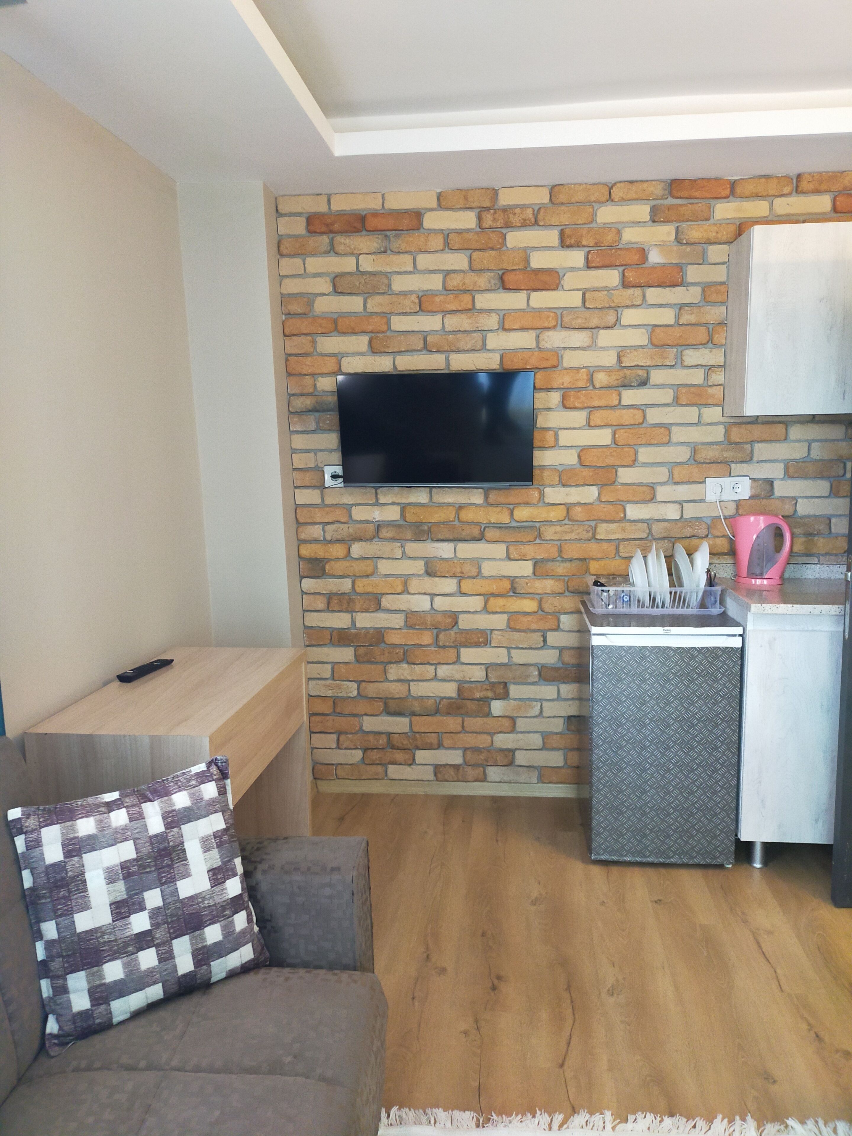Un espacio habitable compacto con una pared de ladrillo, un televisor de pantalla plana montado encima, una pequeña cocina americana con refrigerador, y un sofá con un cojín estampado.
