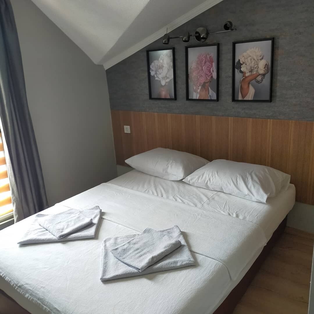 A neatly made bed with white linens and pillows, a wooden headboard, and three framed floral paintings on the wall above.