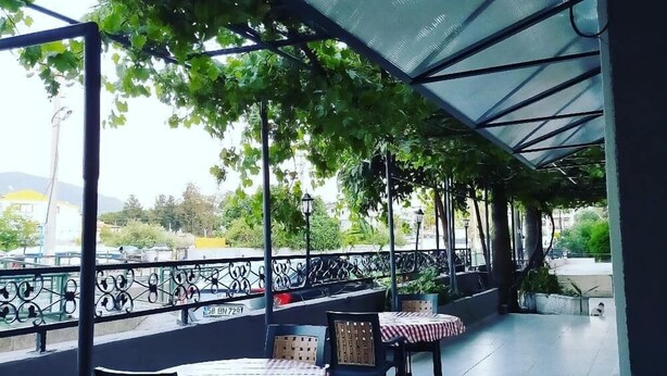 Un café al aire libre con mesas y sillas, cubierto por un techo transparente, rodeado de follaje.