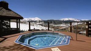Un bain à remous avec vue sur des montagnes enneigées, une terrasse en bois et une rambarde en métal.