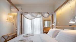 Hotelkamer met een groot bed, witte lakens, een houten hoofdbord, een nachtkastje met een lamp en een groot raam met dunne gordijnen.