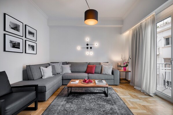 A modern living room with a grey sectional sofa, a black armchair, a wooden coffee table with books and flowers, a patterned rug, and a large window with curtains.