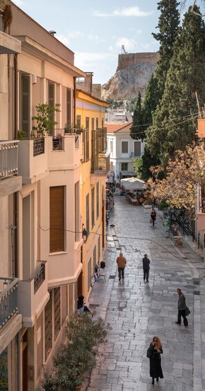 A pedestrian street with buildings on either side, a rocky hill in the background, and a few people walking.