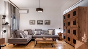 Een moderne woonkamer met een grijze modulaire bank, een houten salontafel en een visgraat parketvloer. Er hangen drie ingelijste foto’s aan de muur, een plafondlamp en een grote houten kast met meerdere deuren.