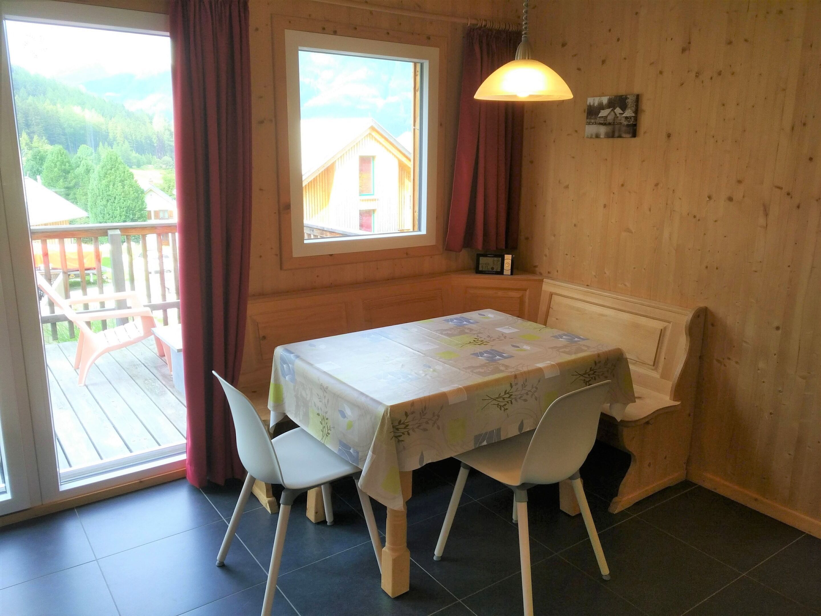 A room with a table and chairs, a window with curtains, a wooden wall, and a view of a house.