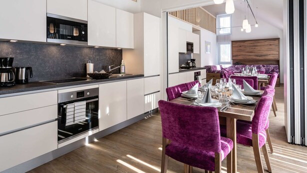 A modern kitchen with white cabinets, a black countertop, and a dining area with purple chairs and a wooden table.