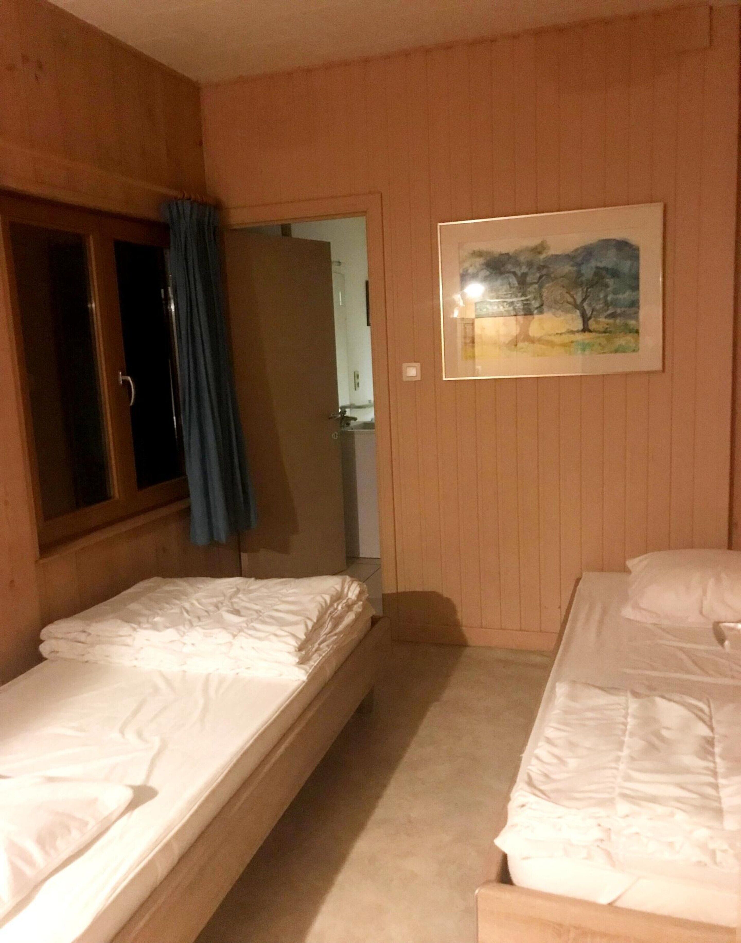 A room with two single beds, wooden walls, a painting, and a door leading to another room.