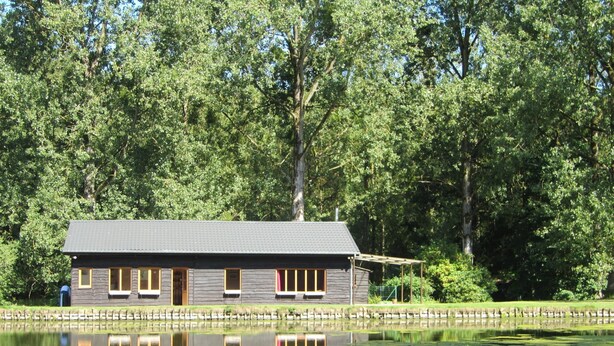 Une cabane au bord d’un lac, dont le reflet se mire dans l’eau, entourée d’arbres.