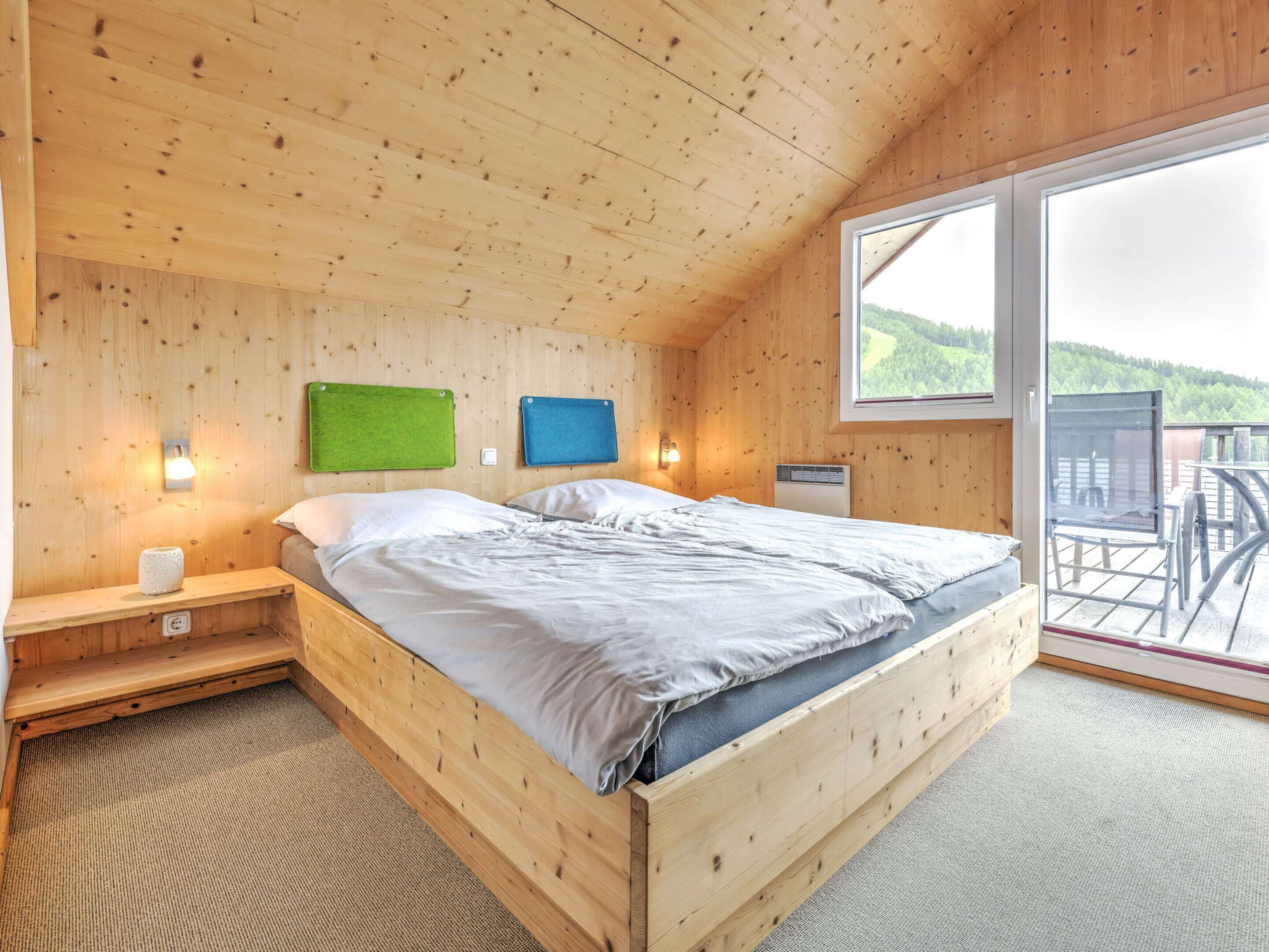 Ein Schlafzimmer einer Holzhütte mit einem Bett, zwei Wandleuchten, einem Fenster und Blick auf einen bewaldeten Bereich.
