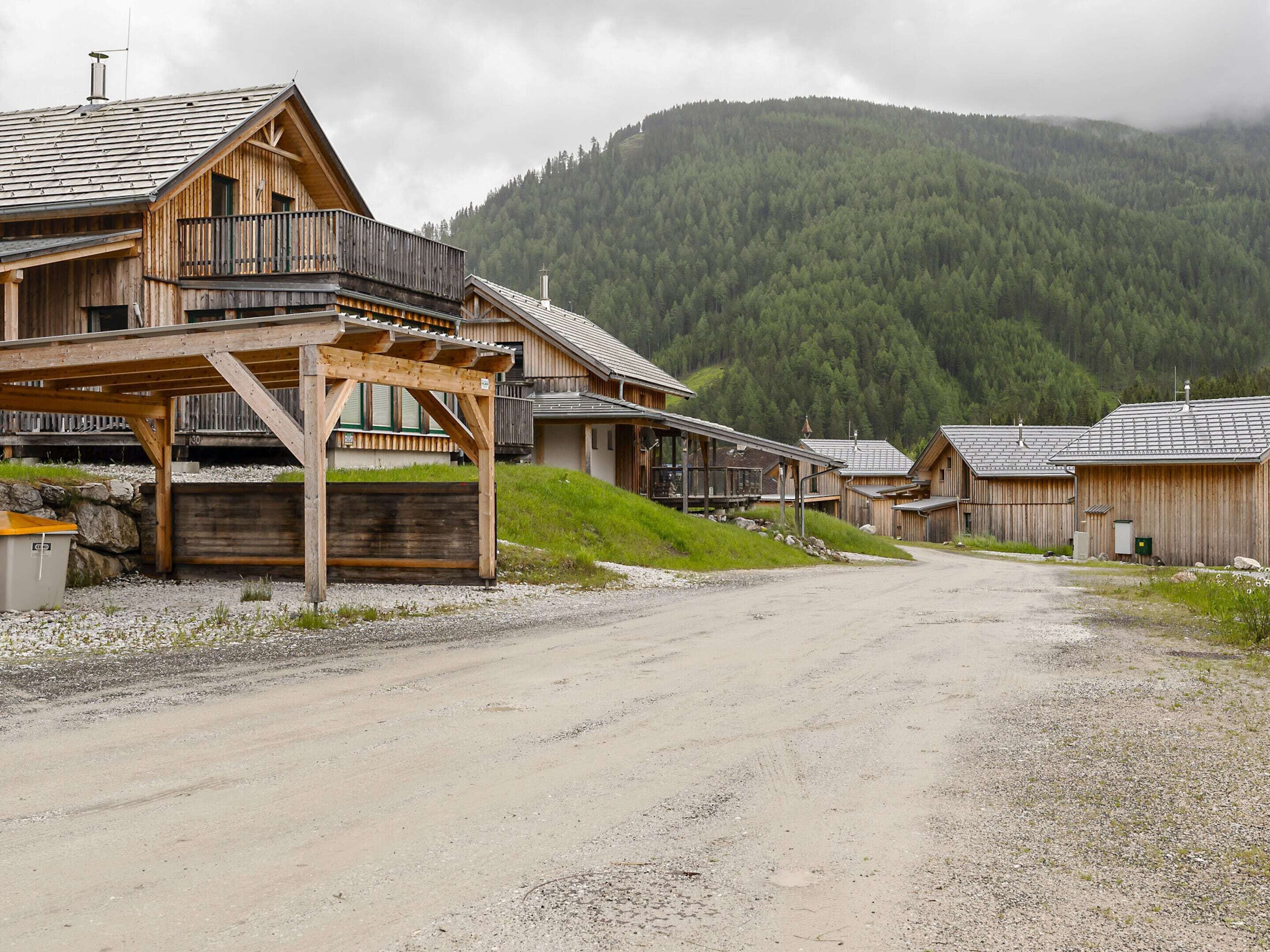 A wooden building with a balcony, a gravel driveway, and a forested mountain in the background.