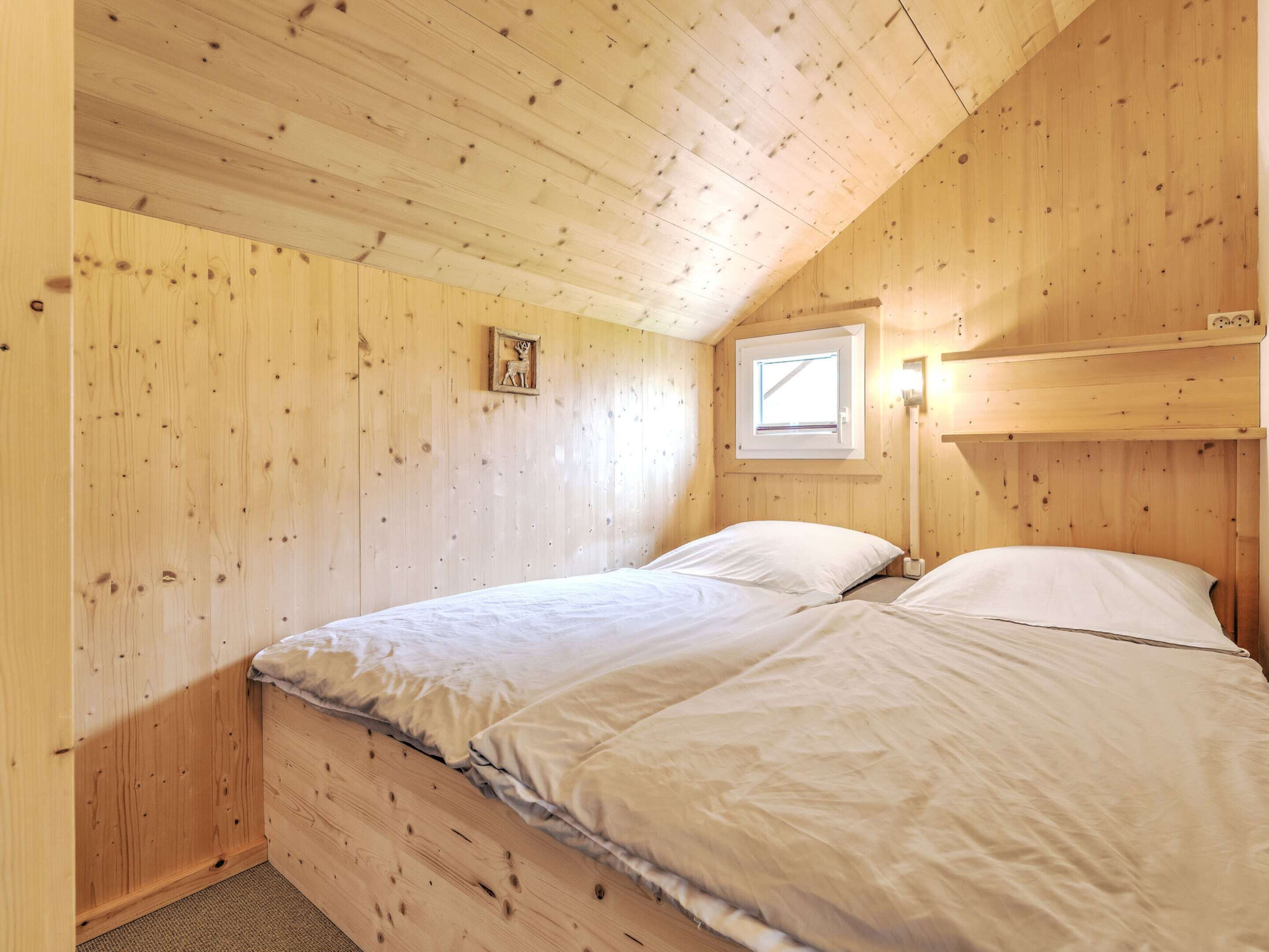 A small, cozy room with wooden walls and a sloped ceiling. There is a bed with white bedding, a small window, and a wall-mounted light fixture.