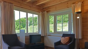 Een gezellige kamer met houten balken, twee fauteuils, een televisie en grote ramen voorzien van gordijnen.