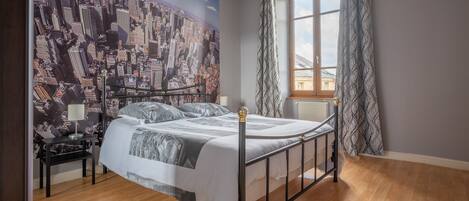 Quarto com mural de paisagem urbana, cama com estrutura metálica, mesa de cabeceira, luminária e janela com cortinas.