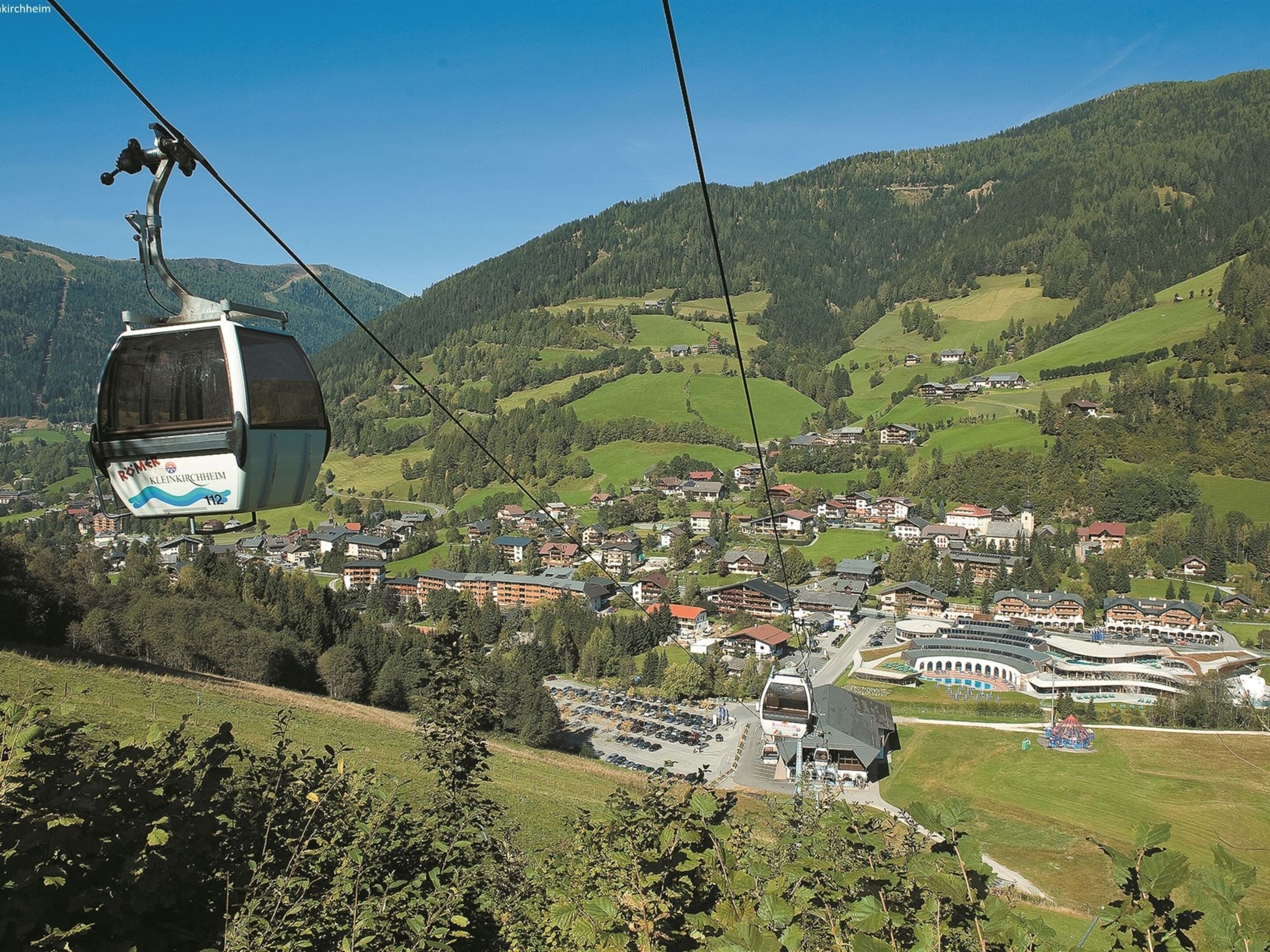 A cable car with "Romex" branding, a town with buildings and a church, and a mountainous backdrop.