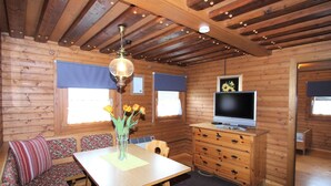 Ein gemütliches Blockhaus-Interieur mit Holzbalkendecke, einem Esstisch mit Blumenvase, einer Couch mit floralen Kissen, einem Fernseher auf einem Holzkasten und einem Fenster mit blauer Jalousie.
