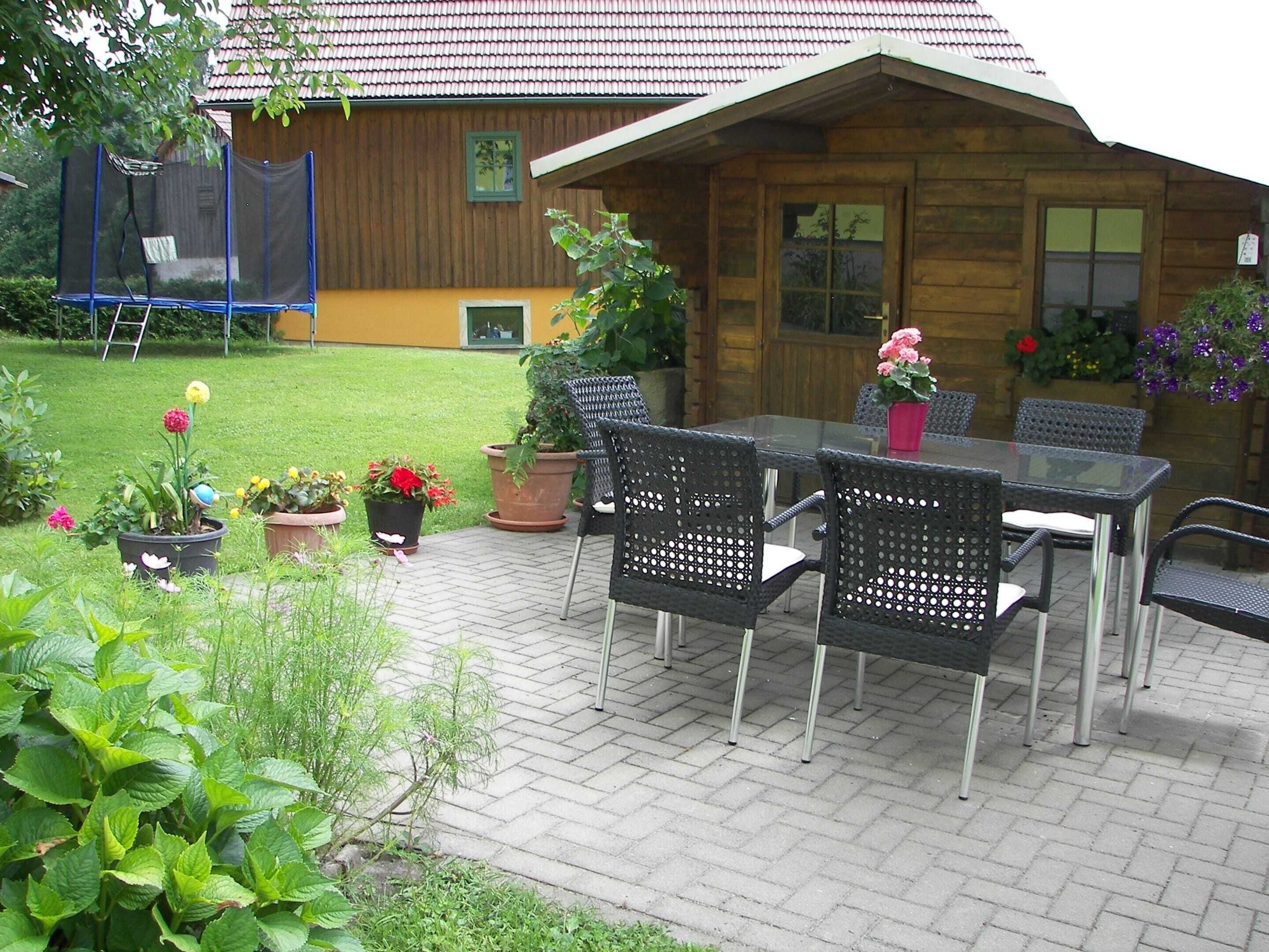 Une maison en bois avec un toit en tuiles, une table en verre entourée de chaises et des plantes en pot dans la cour.