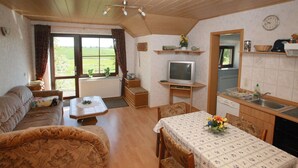 A cozy living room with a sofa, a dining table, and a TV. There's a window with curtains and a view of the outdoors. - Ferienwohnung mit Terrasse in Kropelin (Kroepelin)
